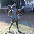 Kleine Nienburgerin Bronzestatue eines jungen Mädchens in Ballerinapose auf gepflastertem Platz vor Geschäften.