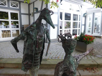 Skulptur "Komm" Zwei abstrahierte Metallskulpturen stehen auf einem gepflasterten Platz vor einem Fenster eines Fachwerkhauses.