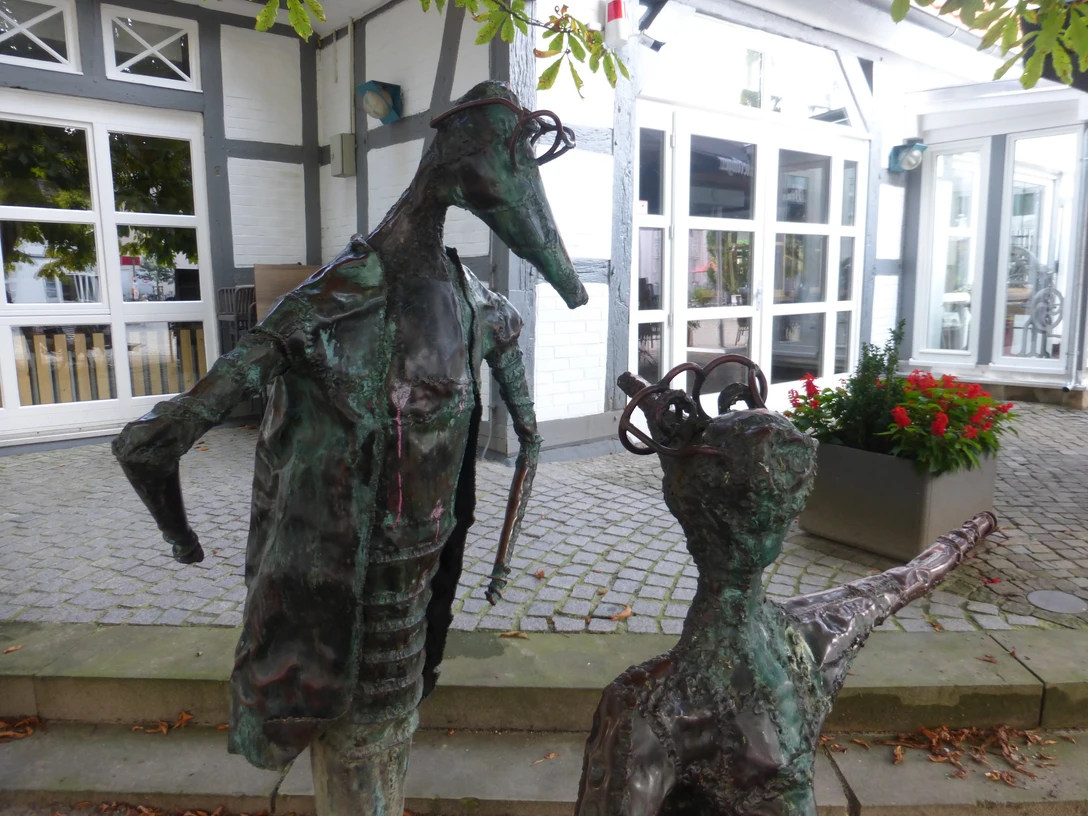 Skulptur "Komm" Zwei abstrahierte Metallskulpturen stehen auf einem gepflasterten Platz vor einem Fenster eines Fachwerkhauses.