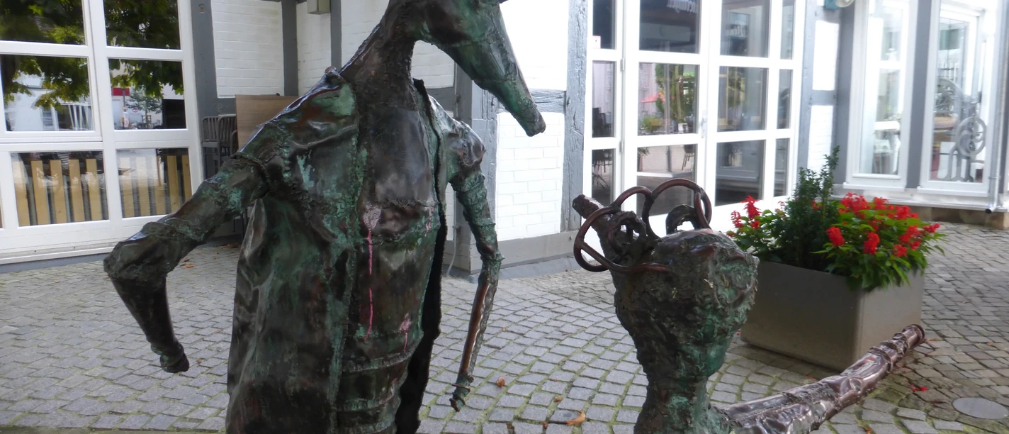 Skulptur "Komm" Zwei abstrahierte Metallskulpturen stehen auf einem gepflasterten Platz vor einem Fenster eines Fachwerkhauses.
