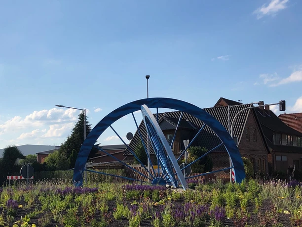 Eine blaue Metallskulptur in Form eines Radkranzes steht im Zentrum eines Kreisverkehrs, umgeben von Blumen.