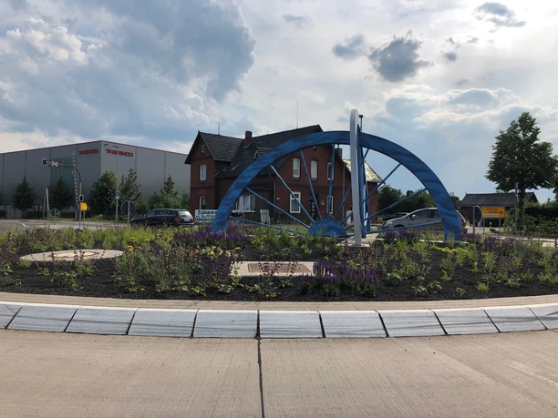 Skulptur Kreisel Hoya Skulptur eines blauen Rades auf einem Kreisverkehr in Hoya, umgeben von Blumen und Industriegebäuden.