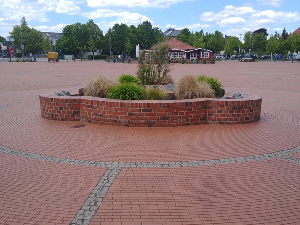 Maskenbrunnen Roter Backsteinbrunnen mit Pflanzen auf gepflastertem Platz, umgeben von Bäumen und einem Kiosk.