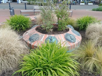 Maskenbrunnen inmitten einer runden Anlage mit Pflanzen und blauer Maske, umgeben von Sitzgelegenheiten.