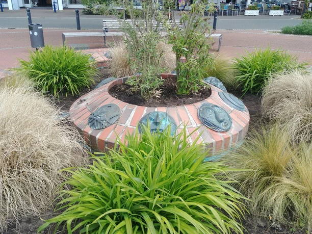 Maskenbrunnen Maskenbrunnen inmitten einer runden Anlage mit Pflanzen und blauer Maske, umgeben von Sitzgelegenheiten.