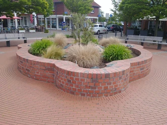 Ein runder Brunnen aus roten Ziegelsteinen mit Gräsern und Pflanzen, umgeben von Sitzmöglichkeiten.