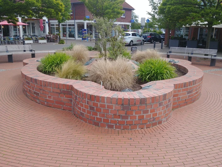 Ein runder Brunnen aus roten Ziegelsteinen mit Gräsern und Pflanzen, umgeben von Sitzmöglichkeiten.