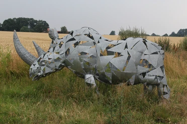 Nashornskulptur aus Metallfragmenten vor einem Getreidefeld und Bäumen, eingehüllt von wilder Vegetation.