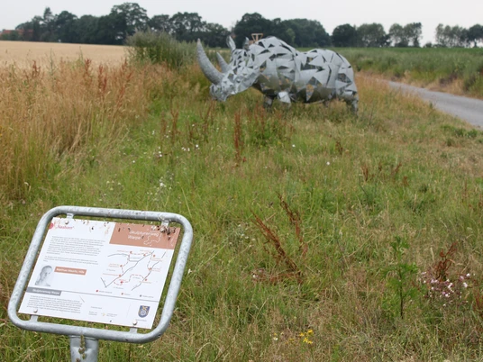 Metallskulptur eines Nashorns in einem grünen Feld nahe einem Weg, mit Infotafel im Vordergrund.