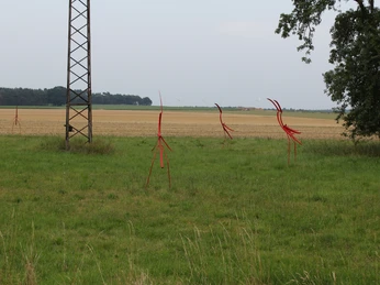 Skulptur Nestbeobachter Rote Metallskulpturen stehen auf einer grünen Wiese inmitten eines flachen Landschaftsbildes.