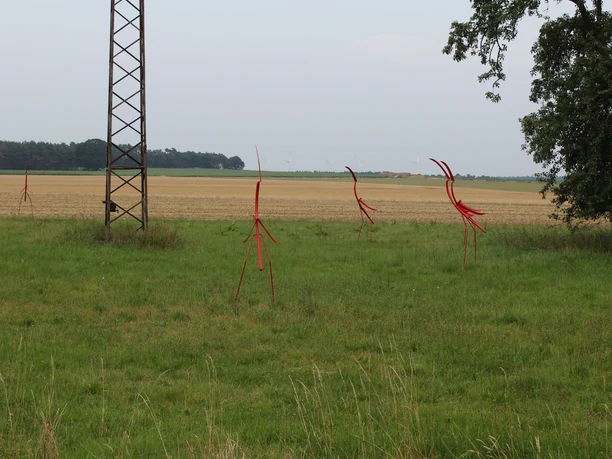 Skulptur Nestbeobachter Rote Metallskulpturen stehen auf einer grünen Wiese inmitten eines flachen Landschaftsbildes.