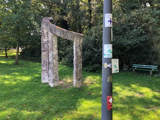 Alter Steinbogen in einem grünen Park mit Bäumen und einer Bank im Hintergrund bei Tageslicht.