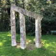 Steinskulptur mit zwei schrägen Säulen und Querbalken, umgeben von Bäumen und grünem Rasen.