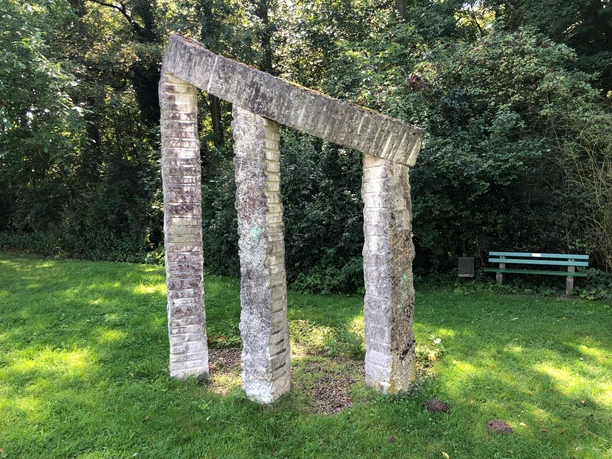 Anfang und Ende Steinskulptur mit zwei schrägen Säulen und Querbalken, umgeben von Bäumen und grünem Rasen.