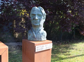 Skulptur Gottlieb Pot d'Or Gottlieb Pot d'Ors Büste steht auf einem metallenen Sockel und wird von Bäumen umrahmt.