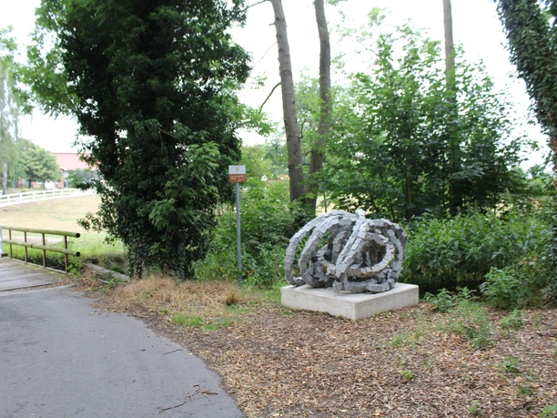 Skulptur Rolle vorwärts aus Stein, umgeben von Bäumen und einem asphaltierten Weg im Vordergrund.