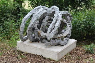 Steinskulptur "Rolle vorwärts" aus granitenen Segmentelementen auf Betonfundament, umgeben von Grün.