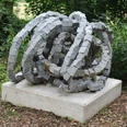 Steinskulptur "Rolle vorwärts" aus granitenen Segmentelementen auf Betonfundament, umgeben von Grün.