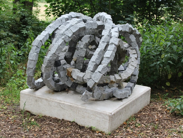 Steinskulptur "Rolle vorwärts" aus granitenen Segmentelementen auf Betonfundament, umgeben von Grün.
