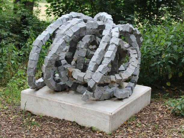 Skulptur Rolle vorwärts Steinskulptur "Rolle vorwärts" aus granitenen Segmentelementen auf Betonfundament, umgeben von Grün.