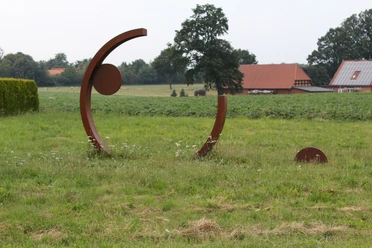 Kunstwerk aus Stahlbögen und Kreisen auf Wiese, umgeben von bäuerlicher Landschaft und Gebäuden.
