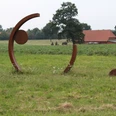 Skulptur Rotationsbewegung Kunstwerk aus Stahlbögen und Kreisen auf Wiese, umgeben von bäuerlicher Landschaft und Gebäuden.