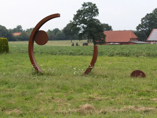 Skulptur Rotationsbewegung Kunstwerk aus Stahlbögen und Kreisen auf Wiese, umgeben von bäuerlicher Landschaft und Gebäuden.