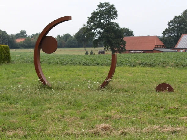Skulptur Rotationsbewegung Kunstwerk aus Stahlbögen und Kreisen auf Wiese, umgeben von bäuerlicher Landschaft und Gebäuden.