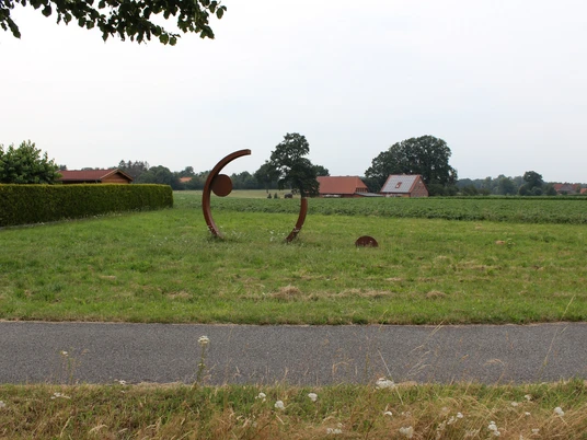 Große, metallene Skulptur in Bewegung darstellend, steht auf einer Wiese, umgeben von Bäumen.