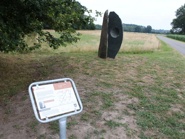 Skulptur "Geöffnete Form" aus Stein mit Loch in ländlicher Landschaft, nahe einem schmalen Weg.