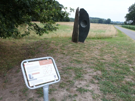Skulptur "Geöffnete Form" Skulptur "Geöffnete Form" aus Stein mit Loch in ländlicher Landschaft, nahe einem schmalen Weg.