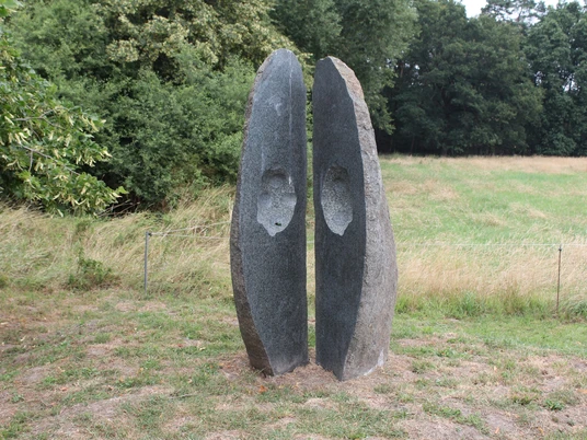 Skulptur aus Stein mit zwei stehend getrennten Halbschalen vor grünem Waldhintergrund.