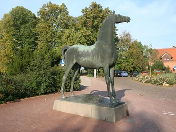 Wiehernder Hengst Bronzestatue eines Hengstes in voller Lebensgröße, erhoben auf einem Sockel, umgeben von herbstlichem Grün.