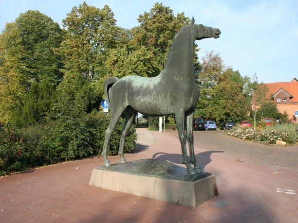 Wiehernder Hengst Bronzestatue eines Hengstes in voller Lebensgröße, erhoben auf einem Sockel, umgeben von herbstlichem Grün.