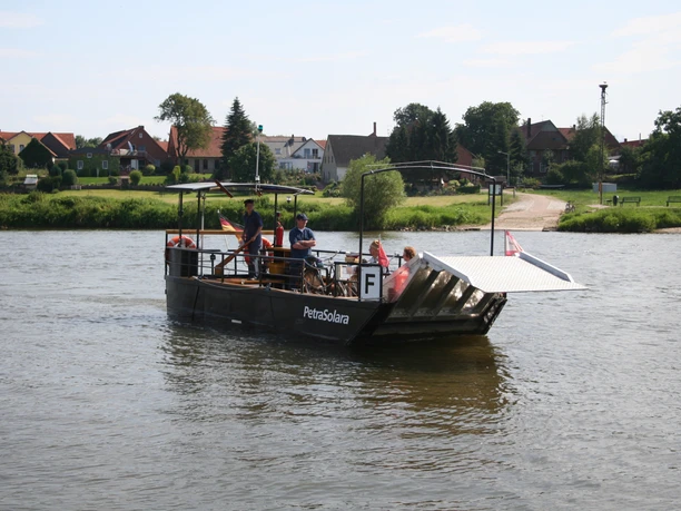 PetraSolara Personen auf einer kleinen Fähre überqueren einen ruhigen Fluss, umgeben von einer ländlichen Landschaft.