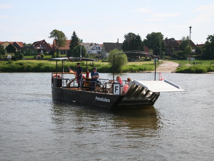 PetraSolara Personen auf einer kleinen Fähre überqueren einen ruhigen Fluss, umgeben von einer ländlichen Landschaft.