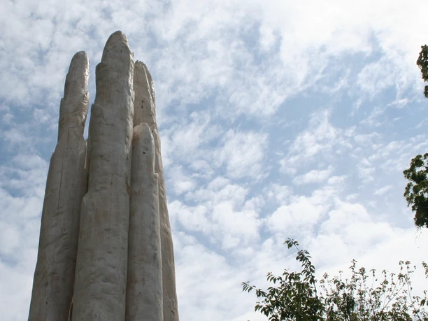 Weiße Spargelskulptur erhebt sich vor einem bewölkten blauen Himmel, umgeben von Bäumen.