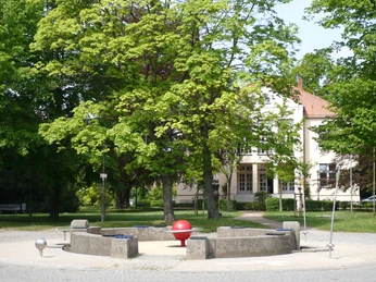 Kreisförmige Sitze mit zentraler rot-oranger Skulptur, umgeben von Bäumen und einem hellen Gebäude.