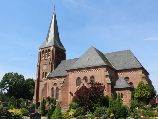 Kirche Lunsen Eine rote Backsteinkirche mit spitzem Turmdach steht vor einem klaren blauen Himmel in grüner Umgebung.