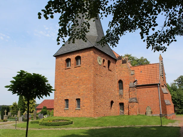 Die Kirche St. Crucis in Hoyerhagen aus rotem Backstein mit Turm steht vor blauem Himmel nahe Bäumen.