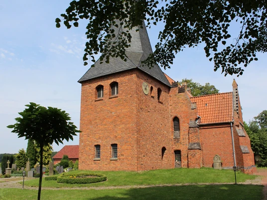 Hoyerhagen St. Crucis Die Kirche St. Crucis in Hoyerhagen aus rotem Backstein mit Turm steht vor blauem Himmel nahe Bäumen.