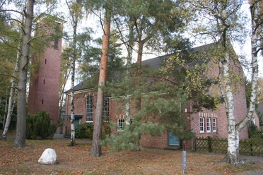Backstein-Kirche im Grünen mit Turm und Fenstern; Bäume im Vordergrund lassen Licht schimmern.