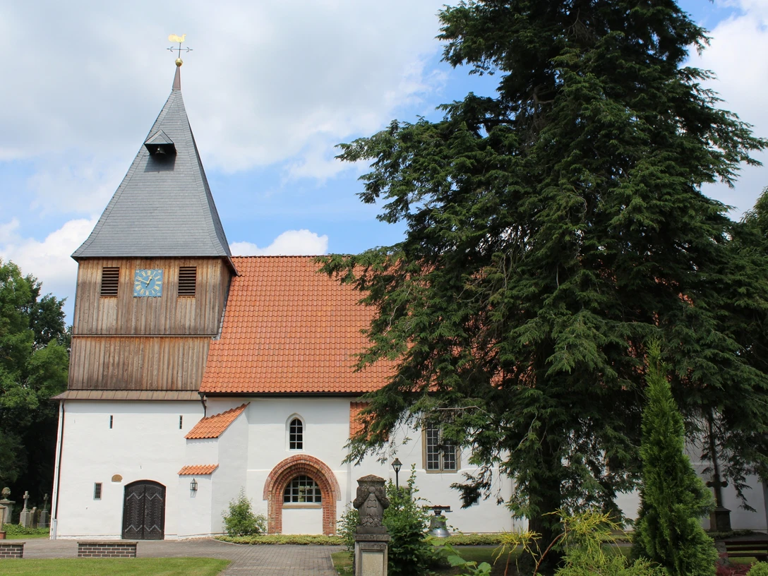 Weißgetünchte Kirche mit rotem Dach und hohem Uhrturm, umgeben von Bäumen unter einem blauen Himmel.