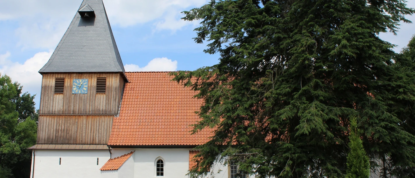 Weißgetünchte Kirche mit rotem Dach und hohem Uhrturm, umgeben von Bäumen unter einem blauen Himmel.