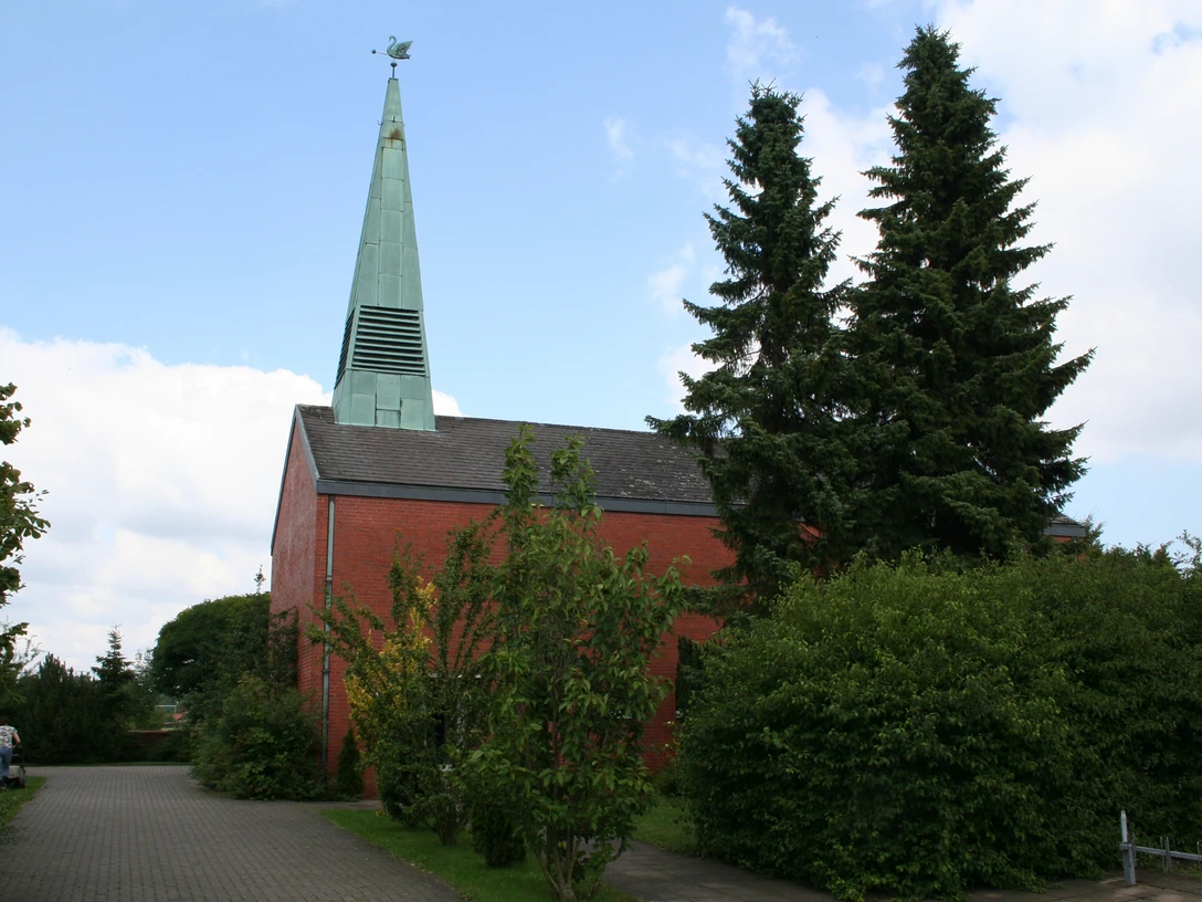 Roter Backsteinbau mit grünem Kirchturm und angrenzenden Bäumen bei bewölktem Himmel.