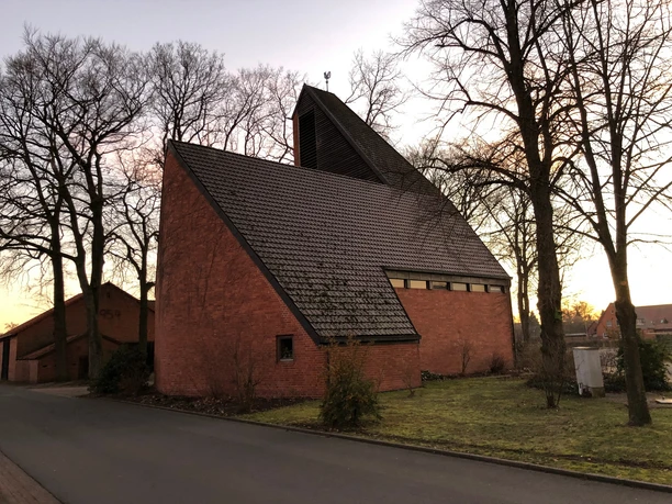 St. Marien Kirche Haßbergen Eine moderne Backsteinkirche mit steilem Dach, umgeben von Bäumen, aufgenommen in der Dämmerung.
