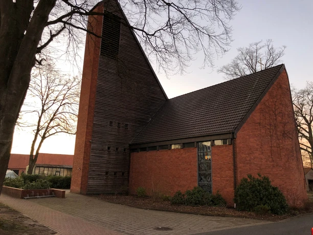Backsteingebäude mit steilem Holzturm und Fenstern, umgeben von kahlen Bäumen und gepflastertem Weg.