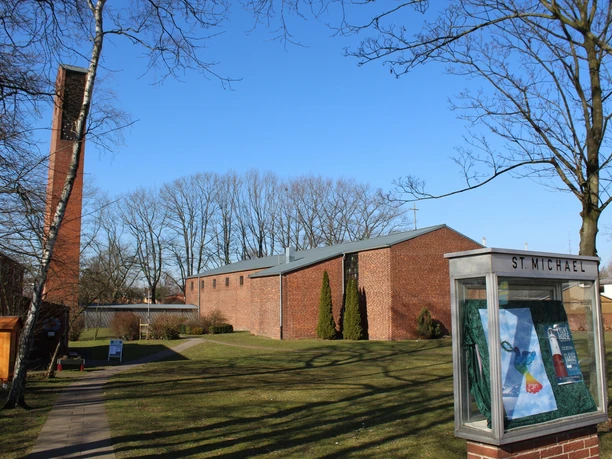 Backsteinkirche mit langem Kirchenschiff und freistehendem Glockenturm, umgeben von Rasen und Bäumen.