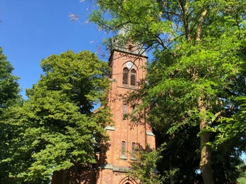 Backsteinkirchturm mit grünem Laub umgeben, unter strahlend blauem Himmel; enthält gotische Elemente.