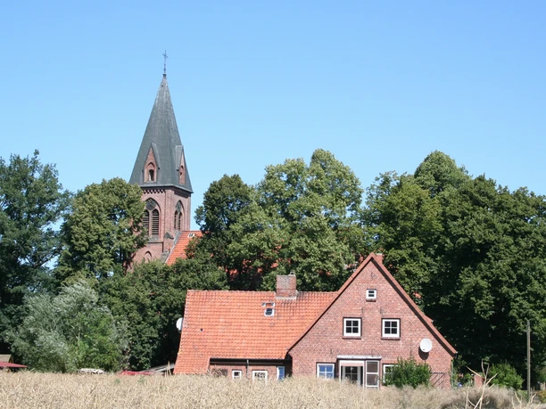 St. Vitus-Kirche Schinna Backsteinkirche mit spitzem Turm hinter einem roten Ziegelhaus umgeben von Bäumen und Feldern.